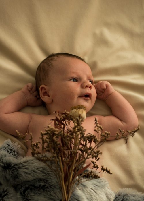 Photographie de bébé Il est allongé sur le dos, les bras relevé avec un bouquet de fleur séchés posées sur lui.